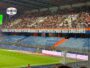 Large crowd of spectators in a stadium stands, with colorful banners along the front and a team logo in the top left corner, indicating a game or event.