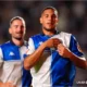 Two soccer players in blue-and-white kits celebrate on the field, the forward adjusting his jersey as a teammate smiles behind him.