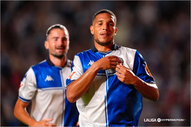 6a9fbbfbe6495 | AllezPaillade.com Two soccer players in blue-and-white kits celebrate on the field, the forward adjusting his jersey as a teammate smiles behind him.