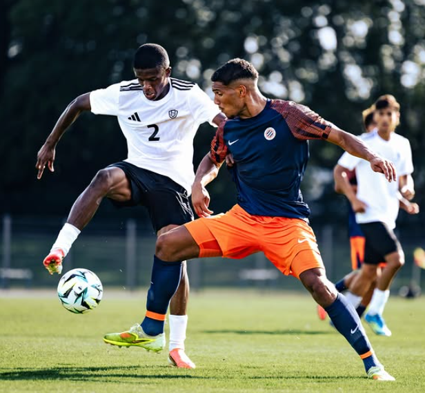 Two soccer players duel for the ball on a sunny field: one in white with number 2, the other in blue and orange, near the ground with the ball between them.