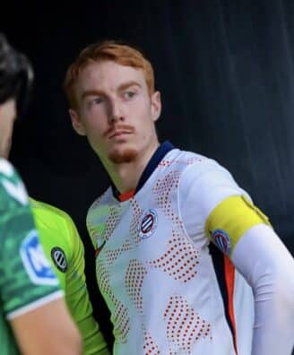 Soccer player in a white jersey with a yellow captain's armband on the left arm, standing with a focused expression.