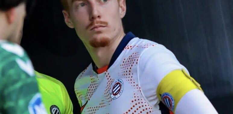 Soccer player in a white jersey with a yellow captain's armband on the left arm, standing with a focused expression.