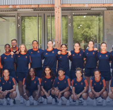 Diverse group of athletes in navy and orange uniforms posing for a team photo outside a building.
