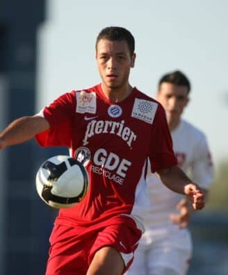 Soccer player in a red kit controls a black-and-white ball during a match, with another player in white in the background.