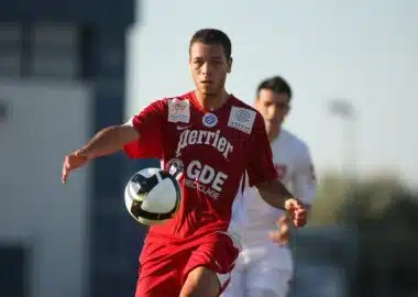 Soccer player in a red kit controls a black-and-white ball during a match, with another player in white in the background.