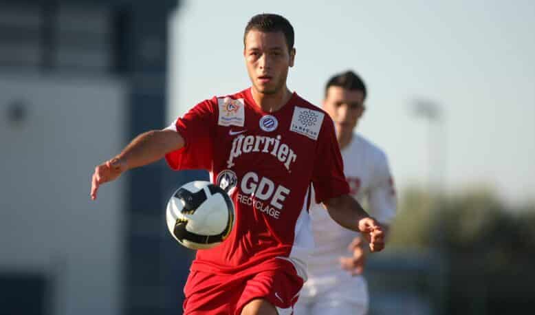 Soccer player in a red kit controls a black-and-white ball during a match, with another player in white in the background.