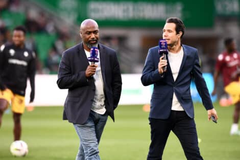 Two male presenters walk on a soccer field, each holding a purple microphone during a live broadcast.