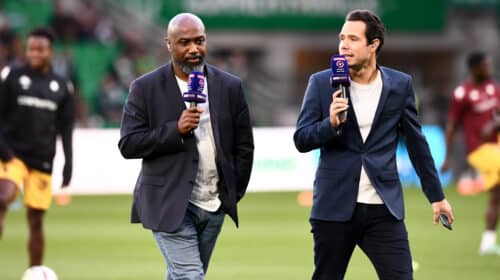 Two male presenters walk on a soccer field, each holding a purple microphone during a live broadcast.