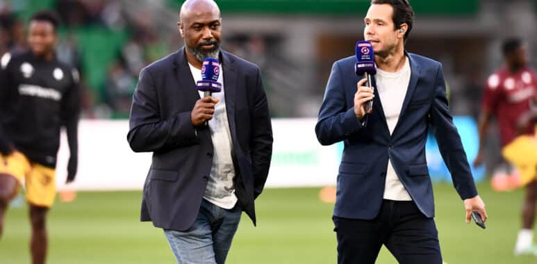 Two male presenters walk on a soccer field, each holding a purple microphone during a live broadcast.