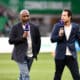 Two male presenters walk on a soccer field, each holding a purple microphone during a live broadcast.