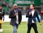 Two male presenters walk on a soccer field, each holding a purple microphone during a live broadcast.