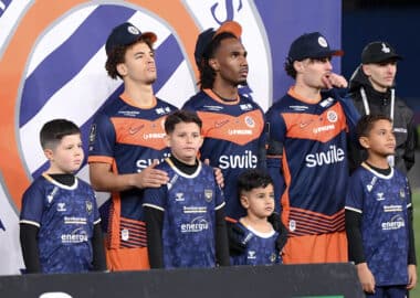 Group of young players in blue and orange jerseys posing on a podium with children in front, backdrop with a circular logo behind them.