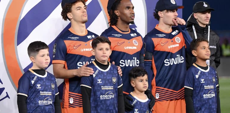 Group of young players in blue and orange jerseys posing on a podium with children in front, backdrop with a circular logo behind them.