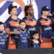 Group of young players in blue and orange jerseys posing on a podium with children in front, backdrop with a circular logo behind them.