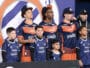 Group of young players in blue and orange jerseys posing on a podium with children in front, backdrop with a circular logo behind them.