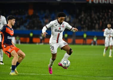 Soccer player in a white kit (number 20) dribbles the ball while a defender in orange pursues him.