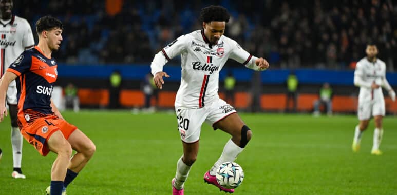 Soccer player in a white kit (number 20) dribbles the ball while a defender in orange pursues him.