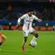 Soccer player in a white kit (number 20) dribbles the ball while a defender in orange pursues him.