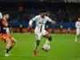 Soccer player in a white kit (number 20) dribbles the ball while a defender in orange pursues him.