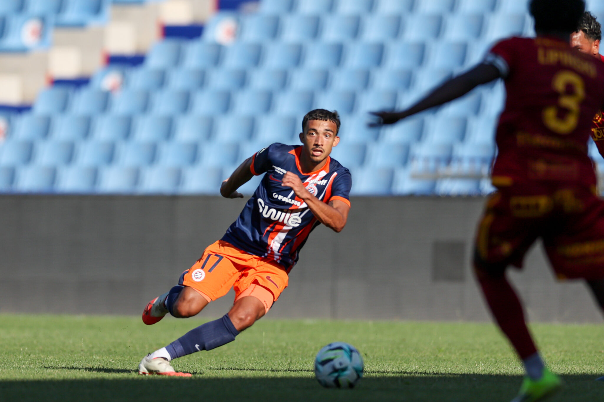 Soccer player in navy and orange kit dribbles the ball on a green field as a rival in maroon closes in.