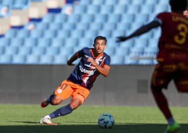 Soccer player in navy and orange kit dribbles the ball on a green field as a rival in maroon closes in.