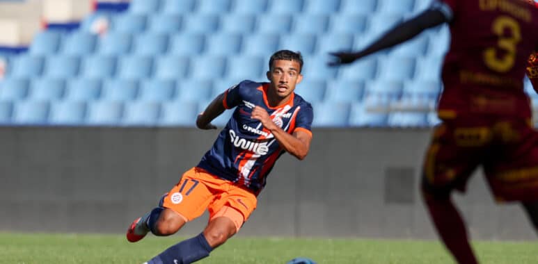 Soccer player in navy and orange kit dribbles the ball on a green field as a rival in maroon closes in.