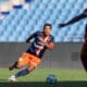 Soccer player in navy and orange kit dribbles the ball on a green field as a rival in maroon closes in.