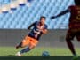 Soccer player in navy and orange kit dribbles the ball on a green field as a rival in maroon closes in.