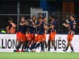 Soccer players in navy and orange celebrate by hugging near the sideline.
