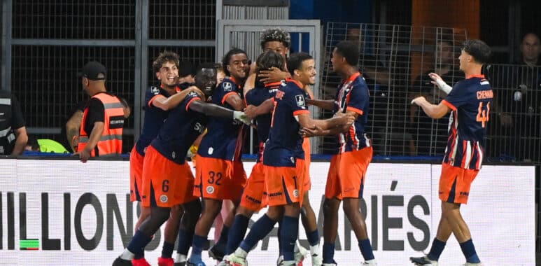 Soccer players in navy and orange celebrate by hugging near the sideline.