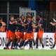 Soccer players in navy and orange celebrate by hugging near the sideline.