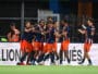 Soccer players in navy and orange celebrate by hugging near the sideline.
