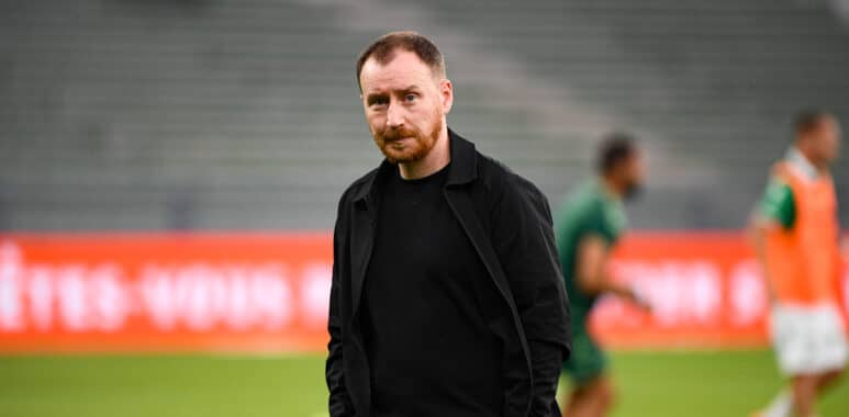 Man in a black jacket standing on a soccer field with blurred players and red advertising boards in the background.