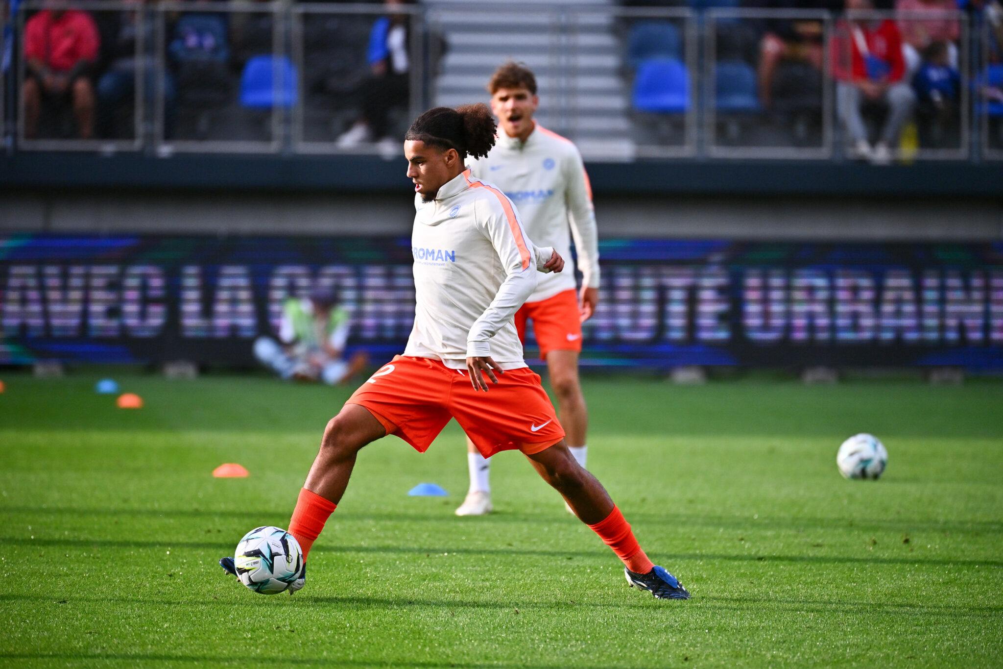 Abderrebi USL Dunkerque v Montpellier HSC - Ligue 2 BKT 20262027 | AllezPaillade.com Soccer player in white jersey and orange shorts dribbles a ball on a green field during practice, with cones and a teammate in the background and stadium seating.