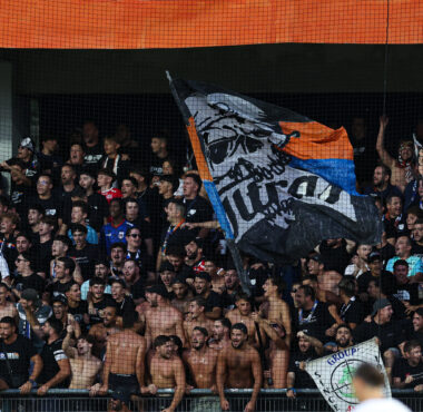 Crowd of excited soccer fans behind protective netting, waving large banners and flags in the stands.