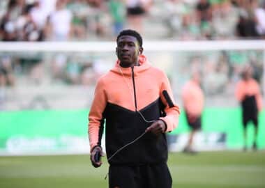 Young man in a peach hoodie and black athletic wear jogging on a soccer field, wearing white earbuds and holding a small device.