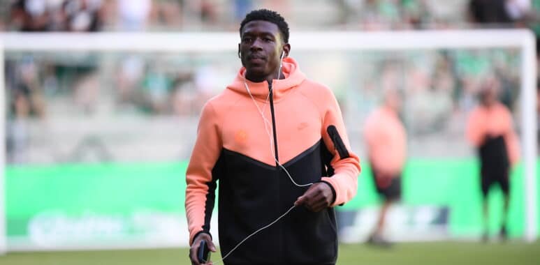 Young man in a peach hoodie and black athletic wear jogging on a soccer field, wearing white earbuds and holding a small device.