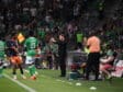 Soccer players in green on the sideline as a coach gestures instructions, with camera crew and a crowded stadium in the background.