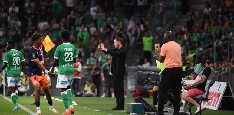 Soccer players in green on the sideline as a coach gestures instructions, with camera crew and a crowded stadium in the background.