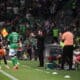 Soccer players in green on the sideline as a coach gestures instructions, with camera crew and a crowded stadium in the background.