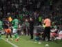 Soccer players in green on the sideline as a coach gestures instructions, with camera crew and a crowded stadium in the background.