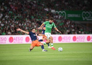 Soccer players challenge for the ball on a green field as a bearded player in a green kit advances while an opposing player slides in from the left.