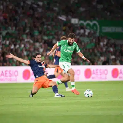 Soccer players challenge for the ball on a green field as a bearded player in a green kit advances while an opposing player slides in from the left.