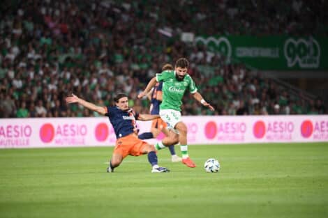 Soccer players challenge for the ball on a green field as a bearded player in a green kit advances while an opposing player slides in from the left.