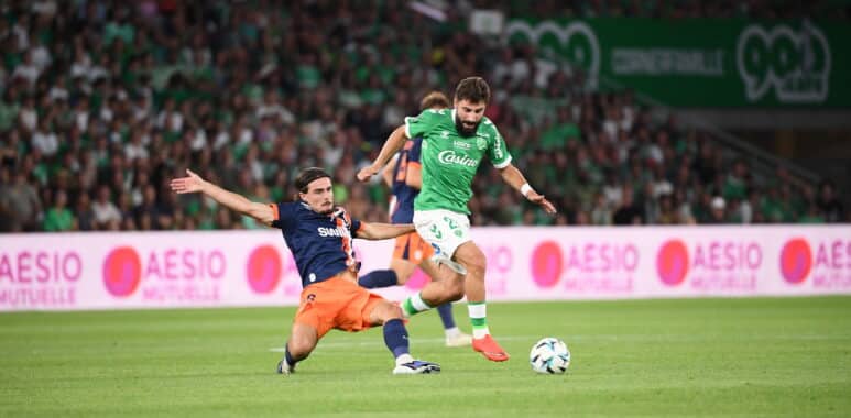 Soccer players challenge for the ball on a green field as a bearded player in a green kit advances while an opposing player slides in from the left.