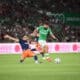 Soccer players challenge for the ball on a green field as a bearded player in a green kit advances while an opposing player slides in from the left.