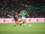 Soccer players challenge for the ball on a green field as a bearded player in a green kit advances while an opposing player slides in from the left.