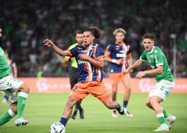 Players in green and navy kits chase the ball on a soccer pitch during a match, with a central player in orange shorts reaching for control.