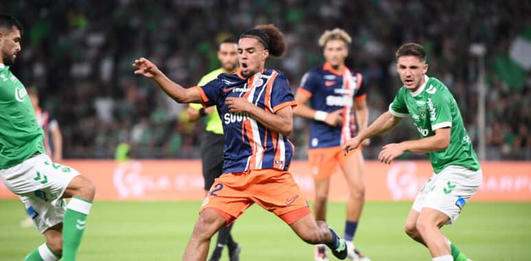 Players in green and navy kits chase the ball on a soccer pitch during a match, with a central player in orange shorts reaching for control.