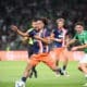 Players in green and navy kits chase the ball on a soccer pitch during a match, with a central player in orange shorts reaching for control.